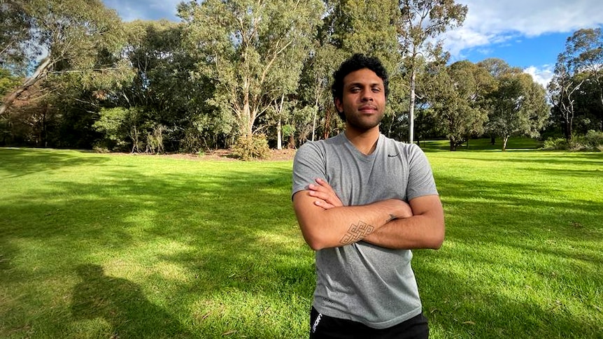 A young man stands in the dappled shade of trees with his arms crossed