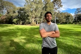 A young man stands in the dappled shade of trees with his arms crossed