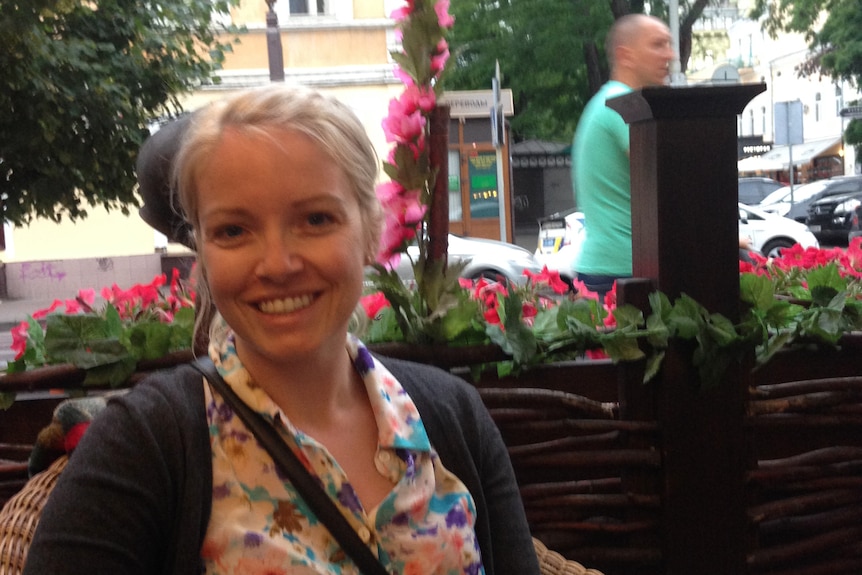 A middle-aged woman with blonde hair smiles at the camera while sitting at an outdoor cafe