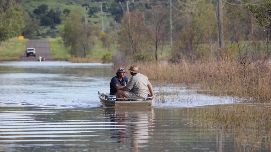 Two men in a tinny, wearing a cap and hat, boating over floodwater, trees, mountain, cars behind.