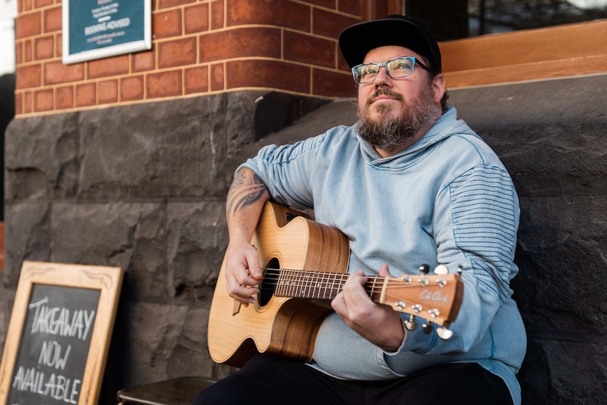 A man sits on a bench in front of an old brick building playing a guitar.