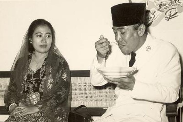 A woman looking at a man wearing Islamic cap holding a plate while eating.