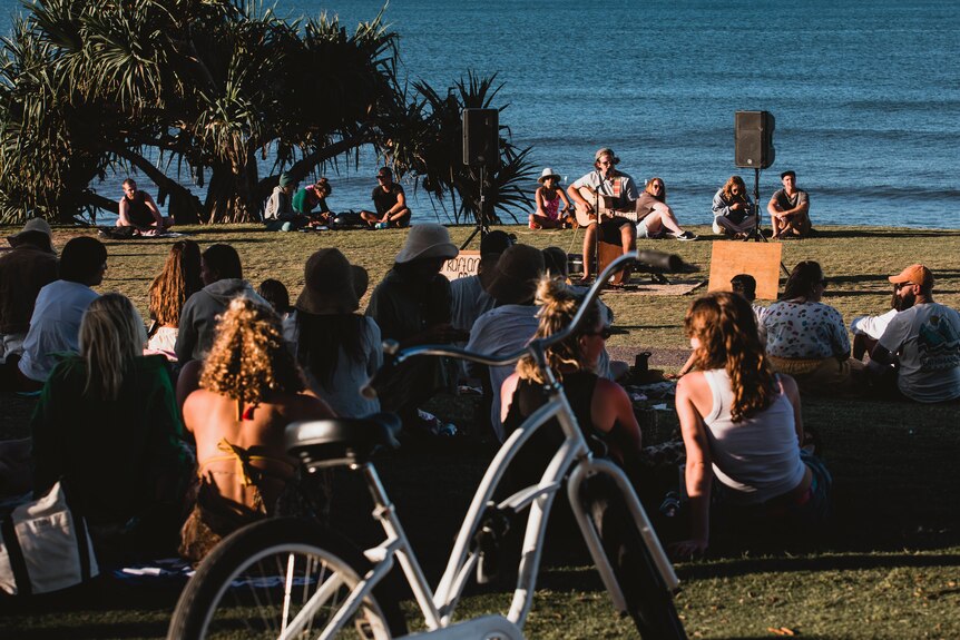 A man plays guitar to a group of people listening in, with the ocean in the background