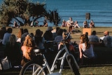 A man plays guitar to a group of people listening in, with the ocean in the background