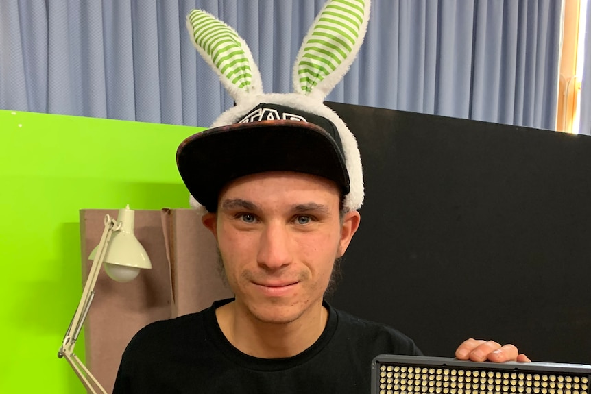 A man wearing rabbit ears stands near a camera 