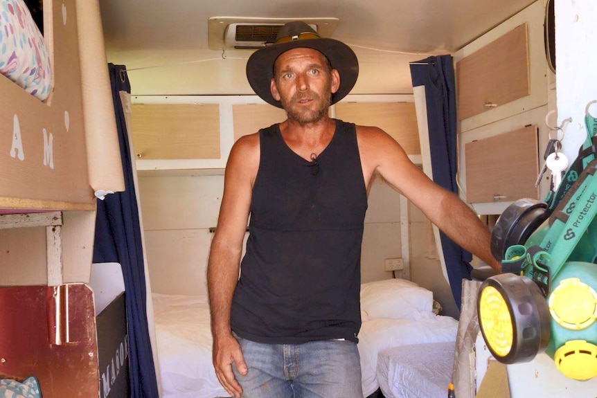 A man in a black singlet and broad-brimmed hat stands inside a converted truck with beds and cupboards.