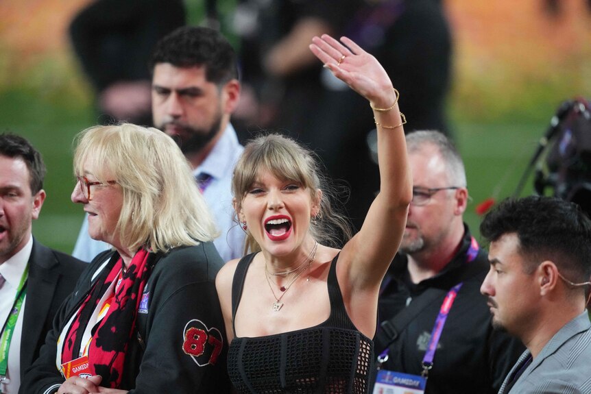 Taylor Swift smiles and waves among a crowd of people on a stadium field.