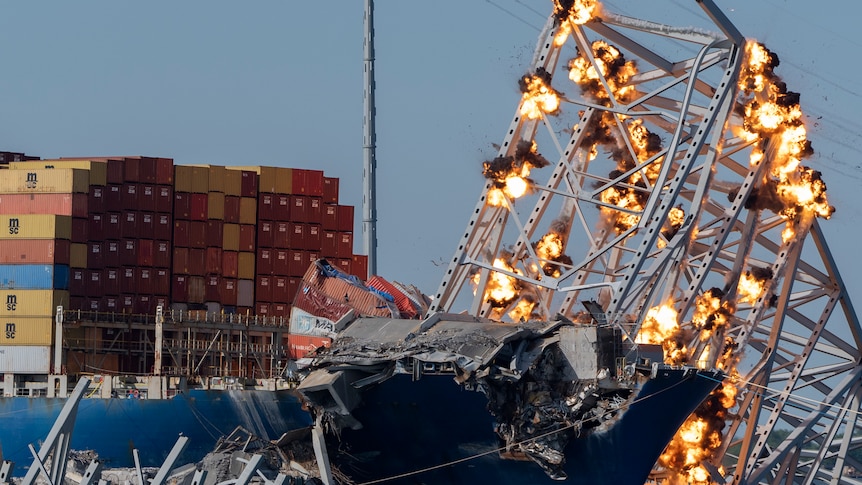 Explosions go off on a damaged section of bridge, which is leaning against a damaged cargo ship