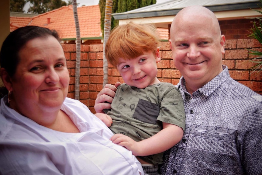 Headshot of a man and a woman holding their young son outside, with a brick wall in the background.