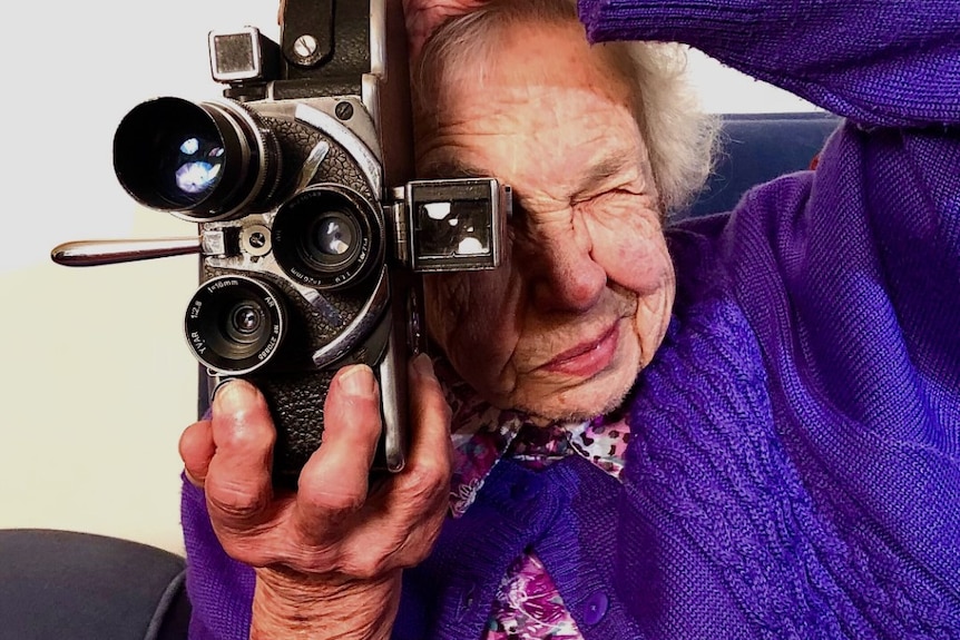 Elderly woman holding old Bolex camera and looking through viewfinder with one eye and other eye screwed up.