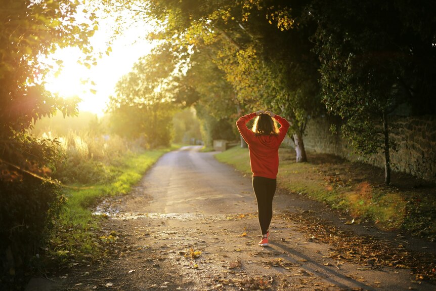 A woman in exercise gear looks down a lonely stretch of road with trees on either side. 