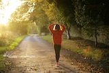 A woman in exercise gear looks down a lonely stretch of road with trees on either side. 