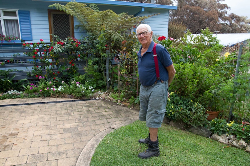 Joe Heaton standing on the grass in front of his flowers and plants and his weatherboard cottage.