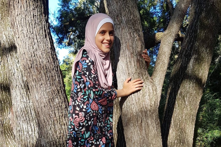 A young teen wearing a hijab smiles as she climbs a tree