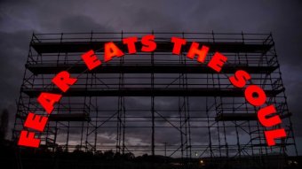 A scaffold against a dark sky with the words 'fear eats the soul', in red, arranged in a semi-circle.