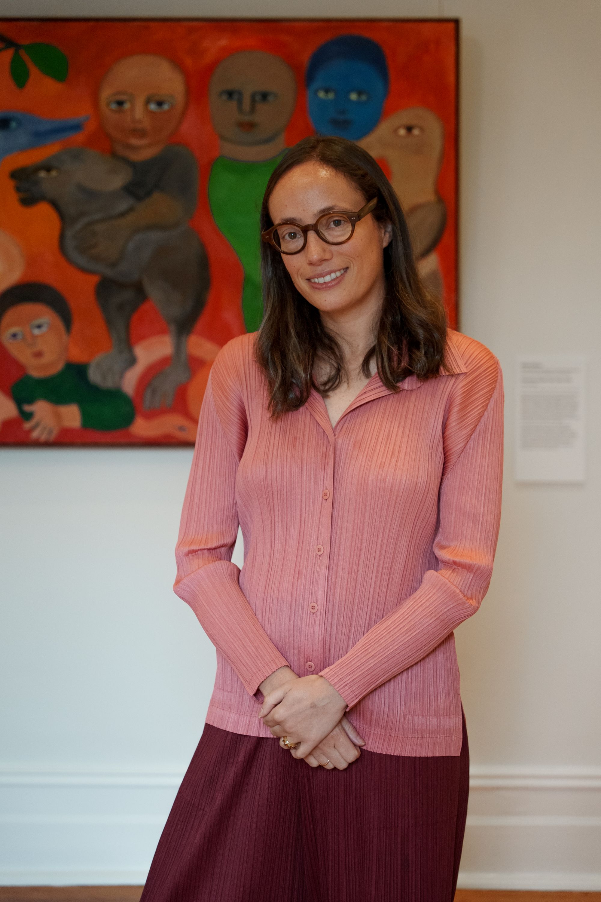 Μια γυναίκα με μακριά καστανά μαλλιά και γυαλιά, φορώντας μια ροζ ζακέτα με κουμπιά, στέκεται μπροστά από έναν πίνακα σε μια γκαλερί.
