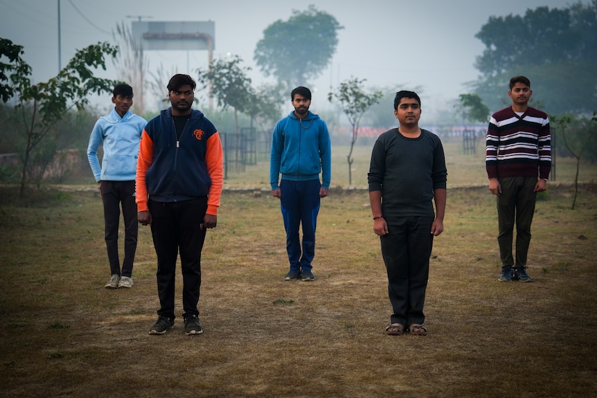 Five men stand with arms by their sides in two rows