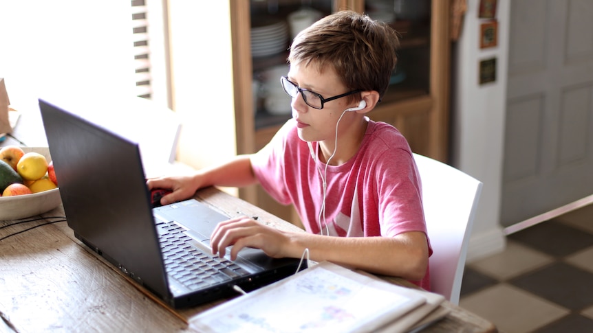 A boy sits in front of a laptop at home, wearing headphones. He looks at the screen. 