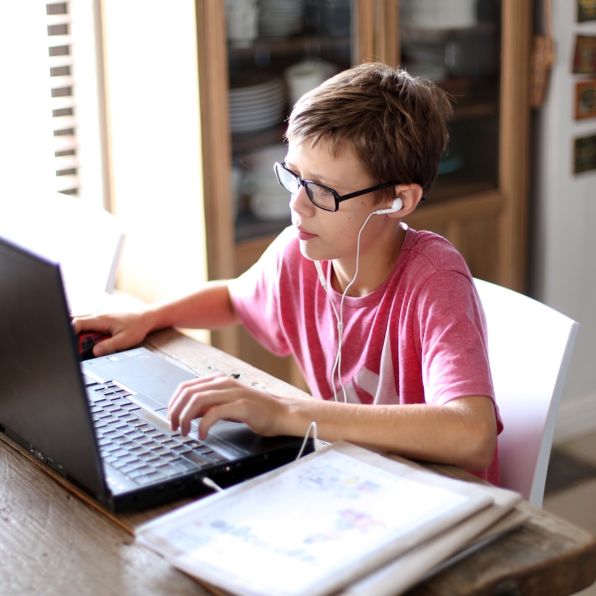 A boy sits in front of a laptop at home, wearing headphones. He looks at the screen. 
