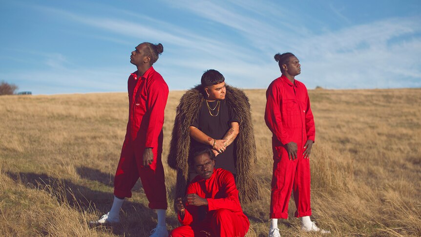 Photo of artist Mojo Juju in a feather coat and three members of Djuki Mala dance group in red jumpsuits posing on grassy plain.