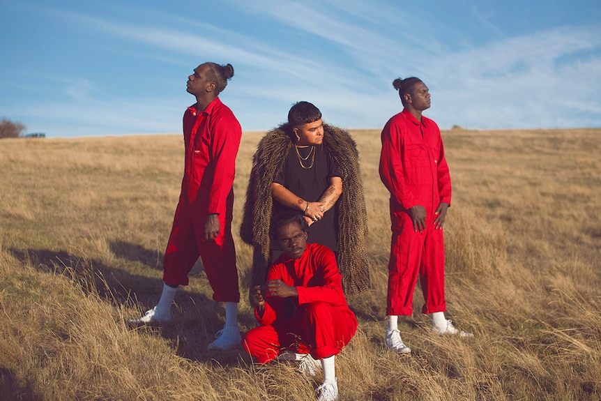 Photo of artist Mojo Juju in a feather coat and three members of Djuki Mala dance group in red jumpsuits posing on grassy plain.