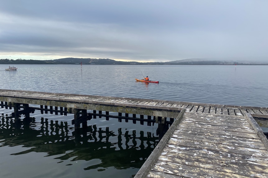A man wearing a high vis jacket is paddling a bright orange kayak across a harbour
