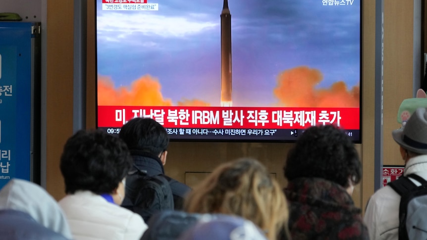 People watch a flatscreen TV showing a rocket launch with red and white korean text underneath.