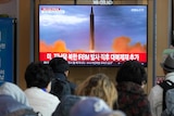 People watch a flatscreen TV showing a rocket launch with red and white korean text underneath.