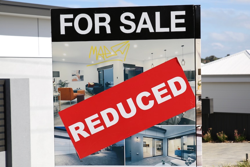 A big red REDUCED sticker is plastered across a For Sale sign in front of a home, alongside graffiti