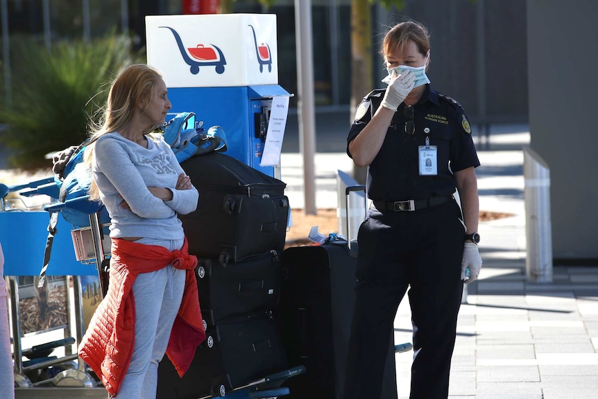 A woman in a tracksuit stands next to a loaded luggage trolley and a Border Force officer wearing a face mask.