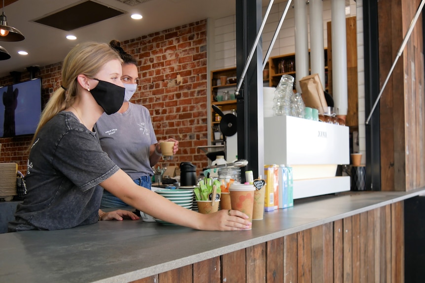 A young girl handing a coffee over a counter with another girl making coffees in the background. Both are wearing masks.