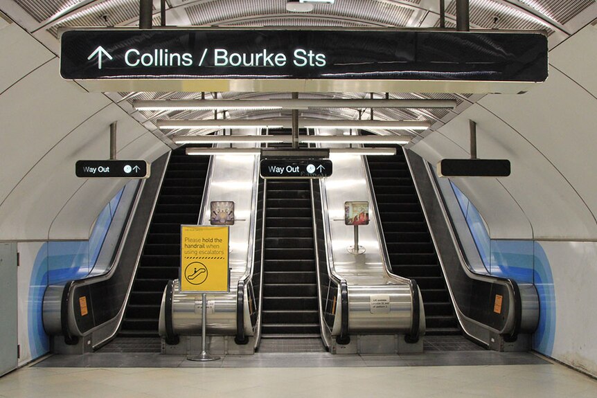 Three escalators in an underground train station, with a sign pointing up to Collins and Bourke streets.