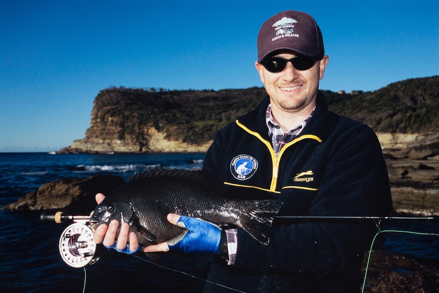 A man holds a fish near the coastline.