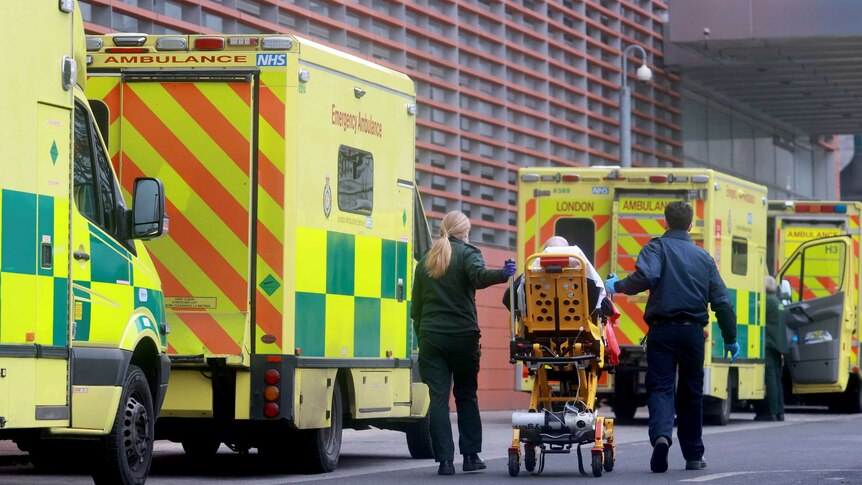 Two paramedics push a patient on a bed past parked ambulances and towards the entrance of the Royal London Hospital.