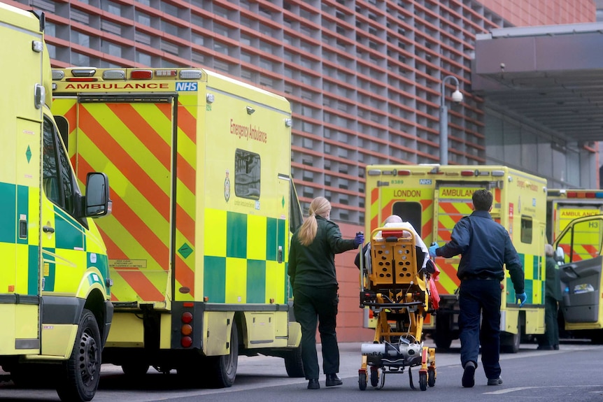 Two paramedics push a patient on a bed past parked ambulances and towards the entrance of the Royal London Hospital.