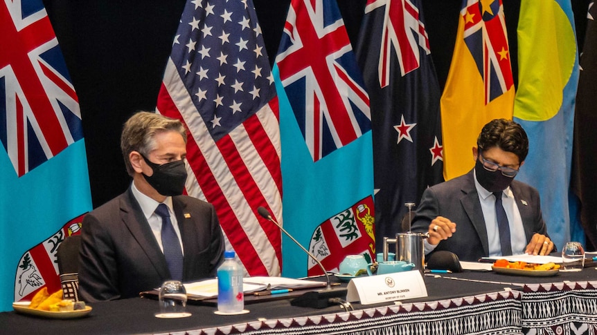 Two suited men wearing masks sitting at a table with multiple flags hanging behind them