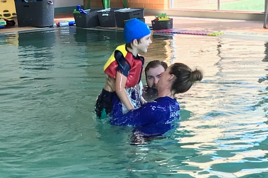 A boy in a swimsuit wearing a floatie being lifted out of the water by a swimming instructor