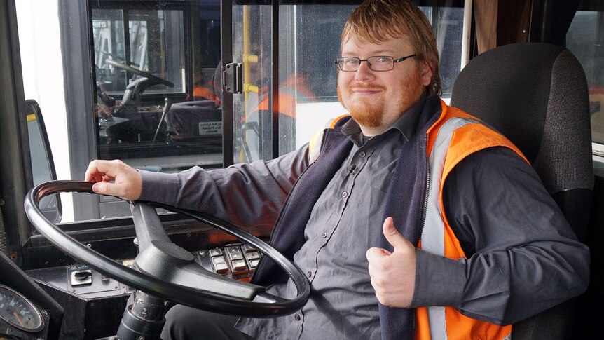Davo Hunter sits in the driver's seat of a bus with one hand on the steering wheel and another giving a thumb's up sign.
