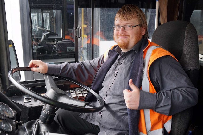 Davo Hunter sits in the driver's seat of a bus with one hand on the steering wheel and another giving a thumb's up sign.