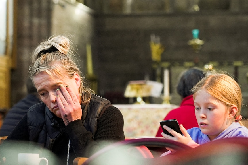 A woman and a child eat and cry while sitting a small church pew inside a dimly lit church.