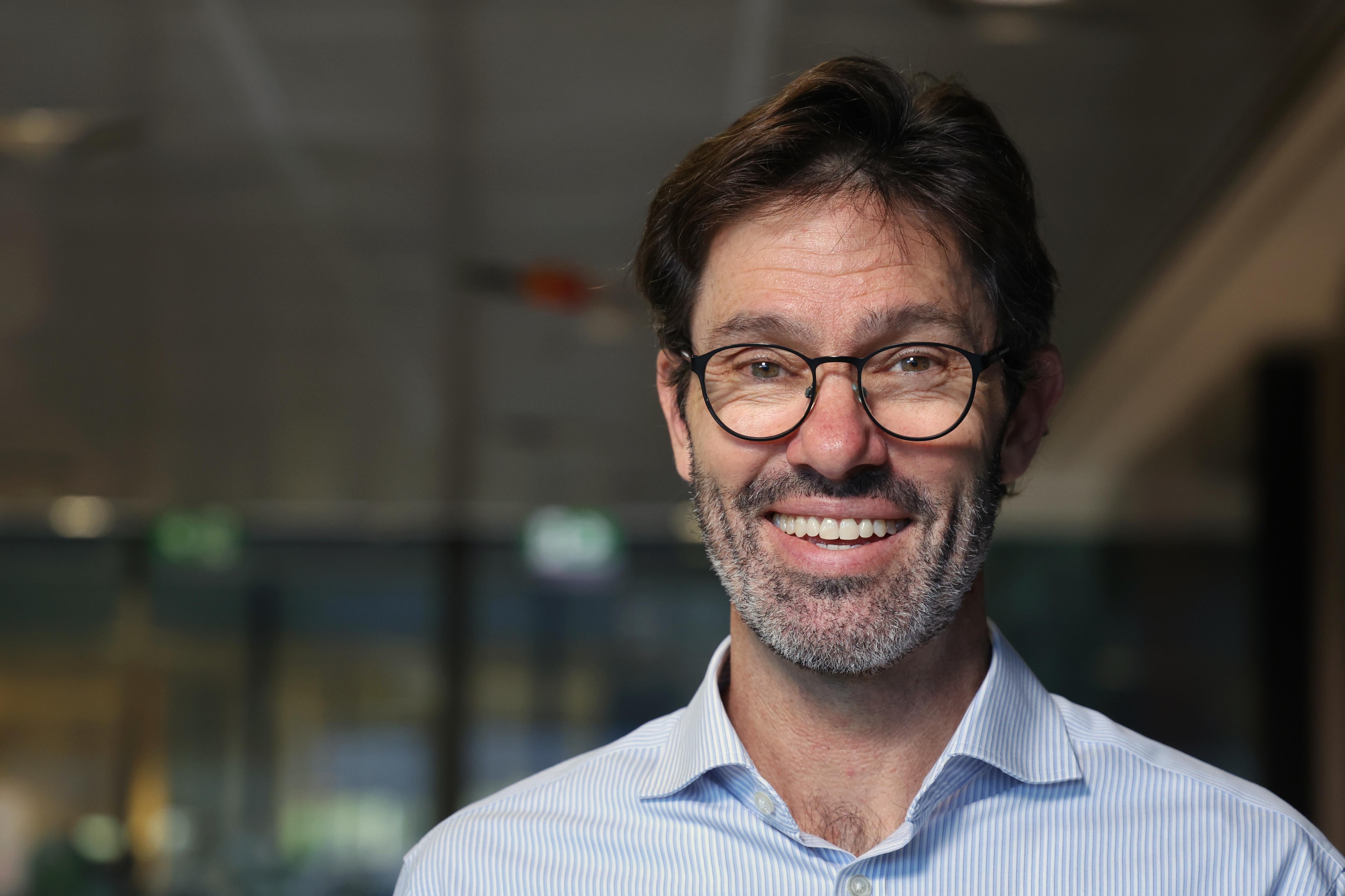 A man in a blue striped collared shirt and glasses smiling into the camera.