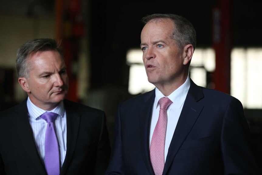 Chris Bowen, wearing a lavender tie and striped shirt, watches Bill Shorten, in a red patterned tie and white shirt, speak.
