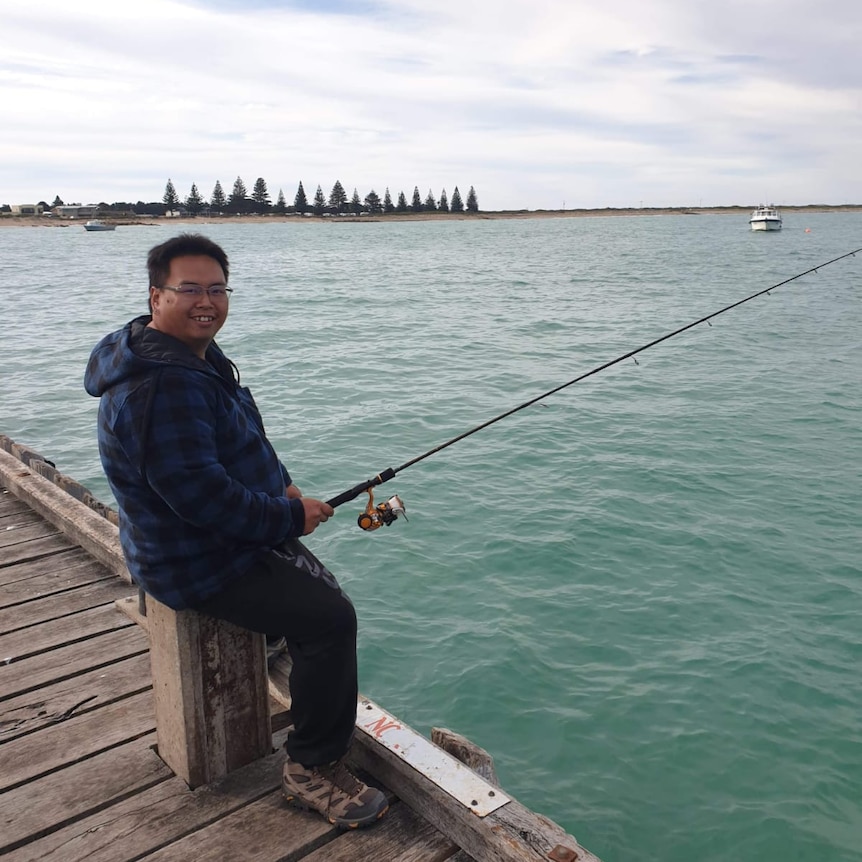 Harrison sitting on a wharf, smiling, holding a fishing rod.