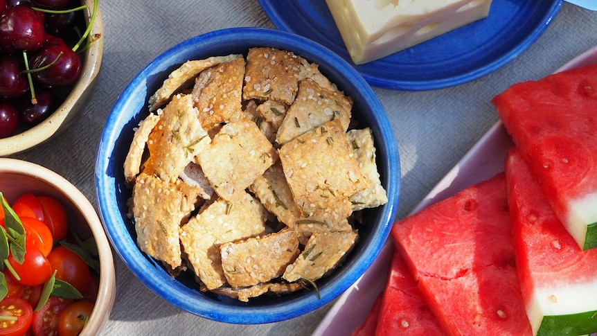 A bowl of sourdough crackers, topped with sea salt, herbs and seeds. Served alongside cheese, fruit, veggies.