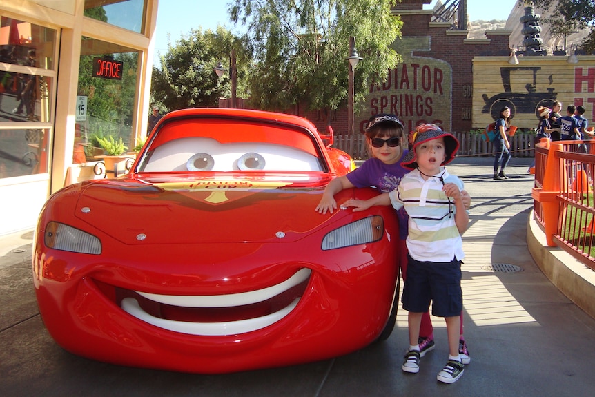 Two kids dressed for day in the sun pose next to a lifesize model of Lightning McQueen.