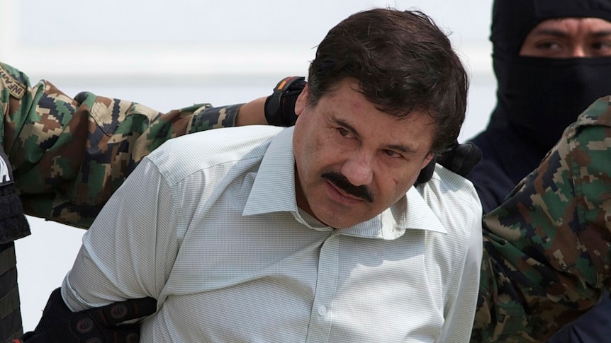 A man with a moustache looks away from the camera as people wearing balaclavas and military fatigues detain him.