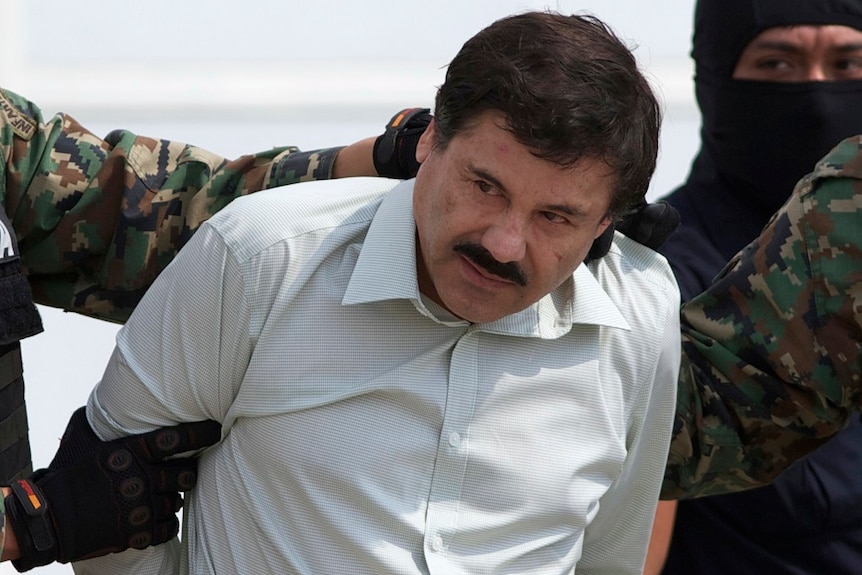 A man with a moustache looks away from the camera as people wearing balaclavas and military fatigues detain him.