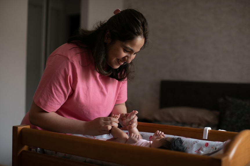 A smiling woman wears pink tee holds newborn baby's feet who is lying on changing table.