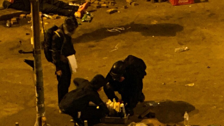 Protesters wearing helmets and protective gear surround a box of molotov cocktails at night.