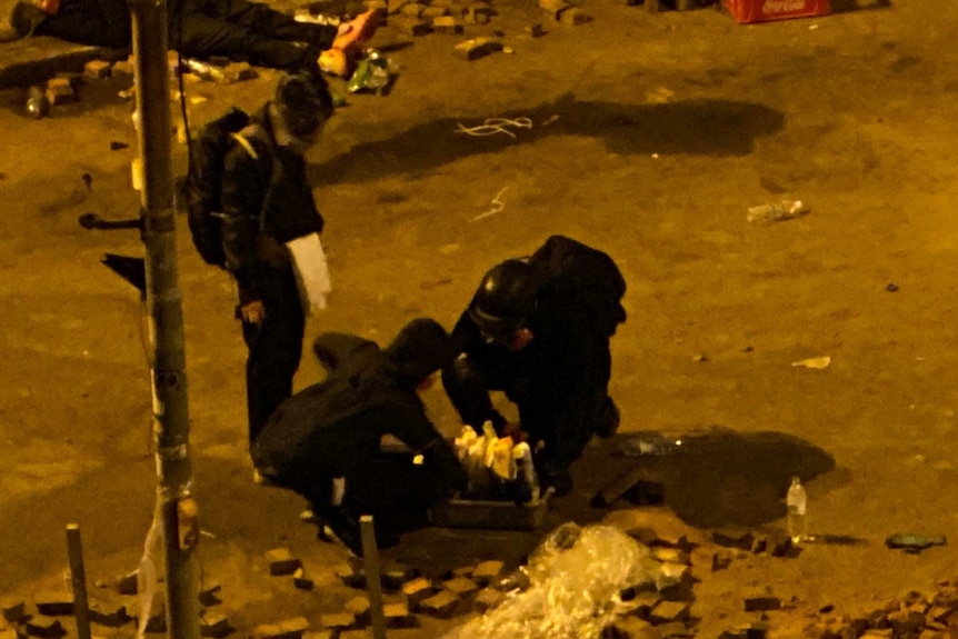 Protesters wearing helmets and protective gear surround a box of molotov cocktails at night.